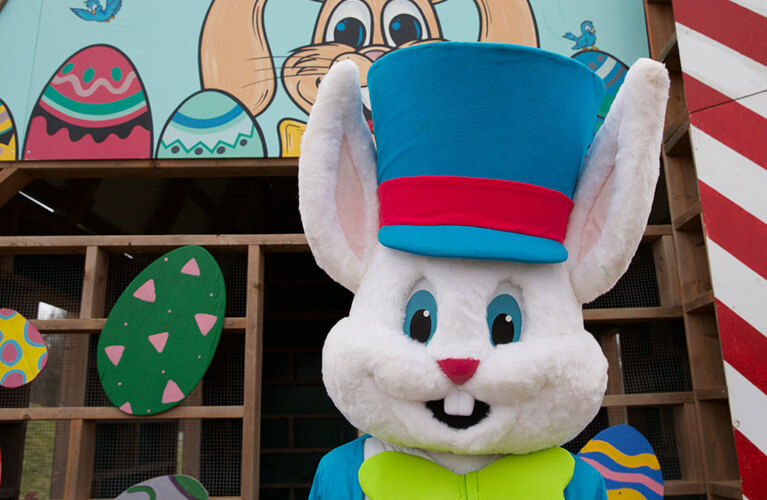 A person in a white Easter Bunny suit with a blue top hat stands before a colorful backdrop of painted eggs and bunny art.