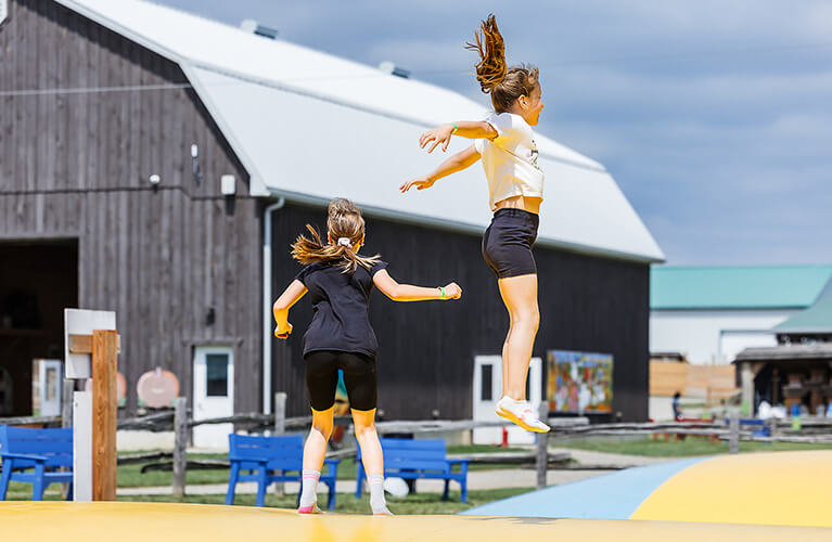 Two girls jump on an outdoor inflatable near a barn, with blue benches and farm buildings in the background.