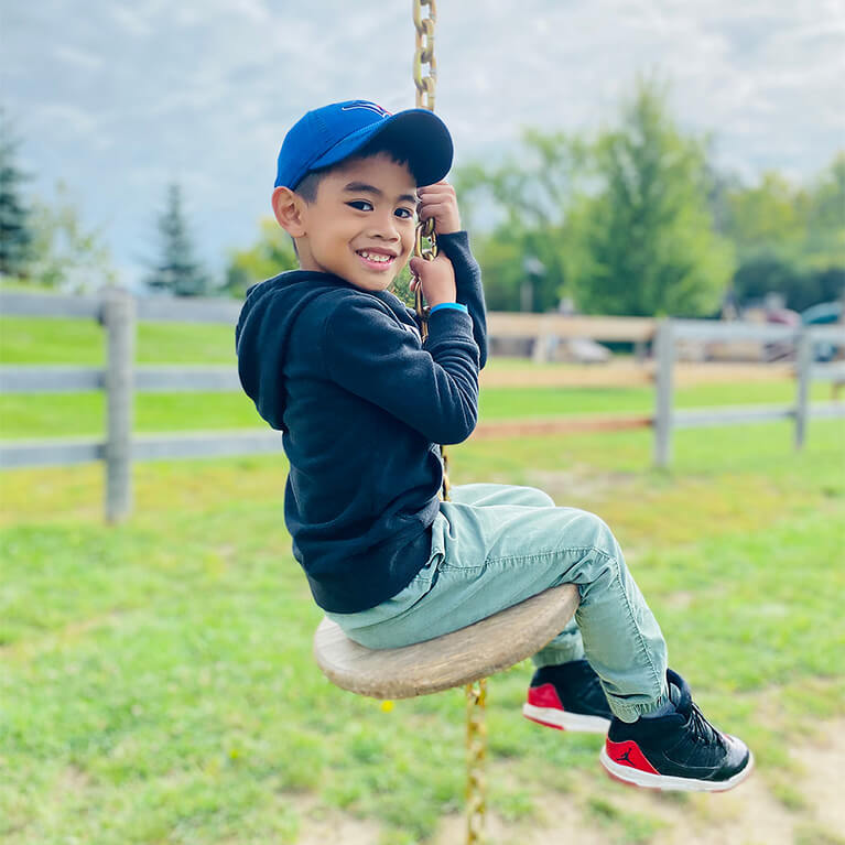 A smiling boy in a blue cap and black hoodie sits on a circular swing outside, surrounded by grass, trees, and a wooden fence.