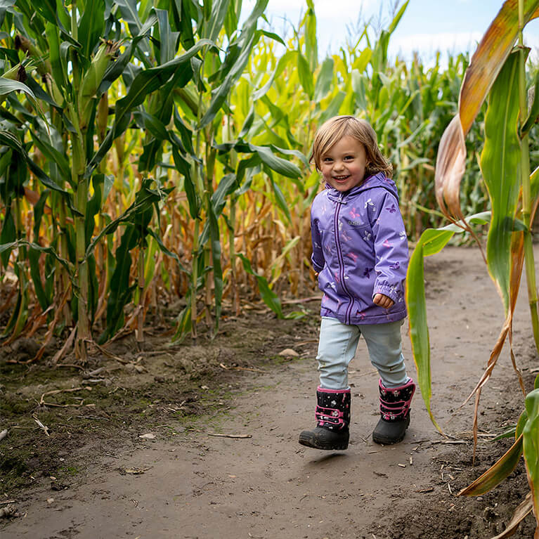 A smiling child in a purple jacket walks along a dirt path between tall green corn stalks on a sunny day.